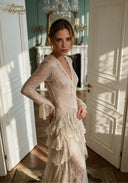 A woman wearing a long-sleeved, sheer beige lace dress with a ruffled tiered skirt, standing in a sunlit room with white paneled doors and a fireplace in the background. The Maison Aphrodite brand logo is visible in the top left corner.