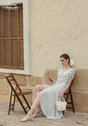 Blue Celeste floral dress with V-neck and side slit, seated and holding woven handbag