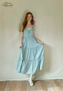 Woman wearing a light blue dress against a plain wall with 'Maison Aphrodite' branding.