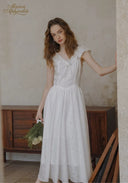 Woman in a white dress holding flowers indoors with a wooden cabinet in the background.