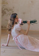 Light pink floral tea dress with puff sleeves and tiered skirt; seated holding a bouquet.