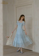 Woman in a light blue dress standing in a room with wooden floor and white walls.