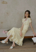 Romance dress in pale yellow with square neckline, puff sleeves, and ruffled hem, seated with woven bag