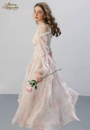 Woman in a long, light pink dress holding a flower against a plain background with 'Maison Aphrodite' branding.