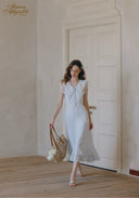 Woman in a white dress holding a straw bag in a room with wooden flooring and a white door.